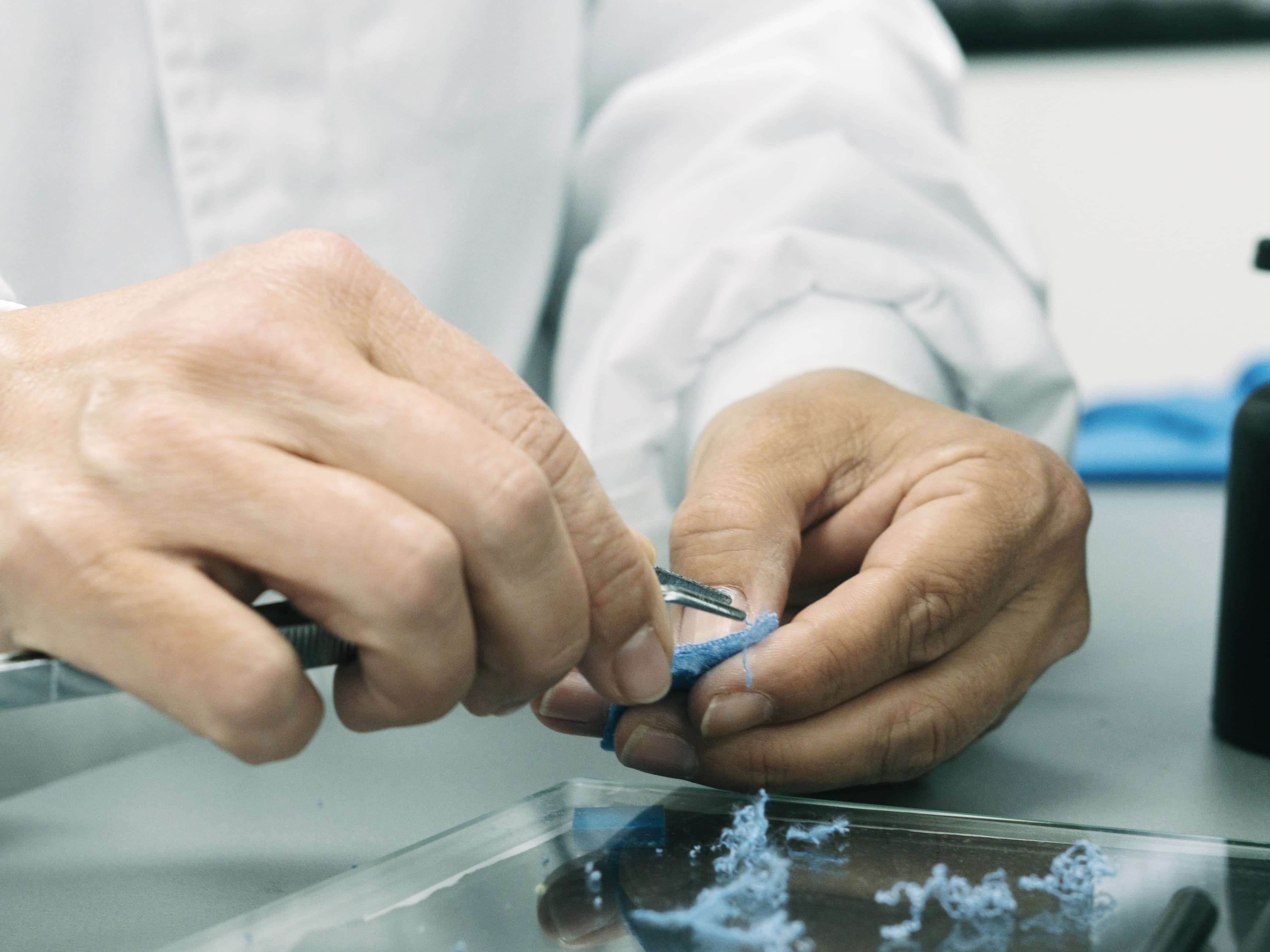 Hands in lab coats inspecting a blue fabric sample with tweezers over a glass surface